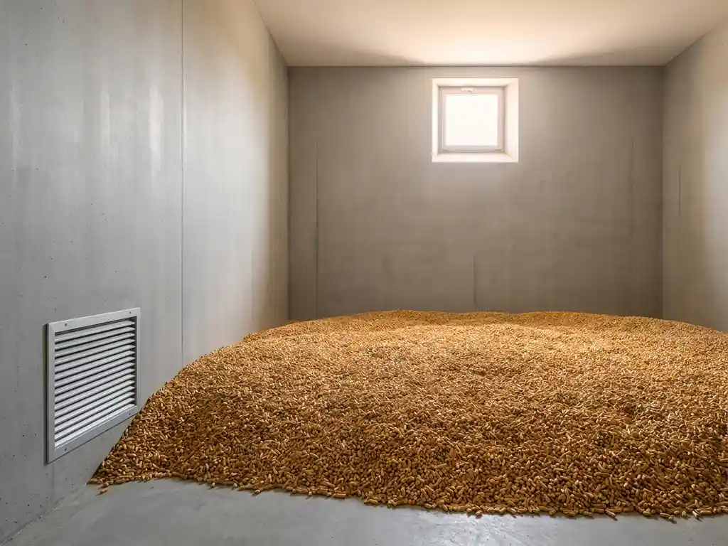 Holzpelletlager mit glattem Betonboden, goldfarbenem Pelletvorrat und Lüftungsgitter an der Wand im minimalistischen Raum.